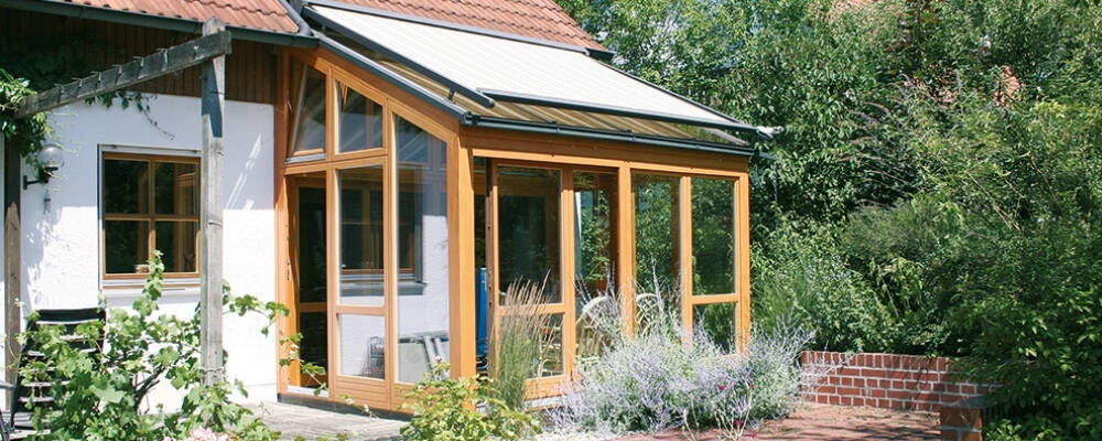 Holz-Wintergarten mit Glasdach und Sonnenschutz am Haus.